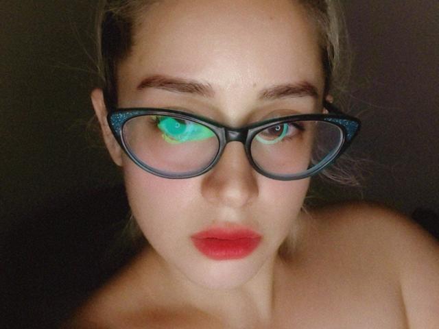 Alexia Morgan, con gafas, reflejo de ojos verde y azul, labios rojos, hombros desnudos, piel clara y cabello rubio, con una expresión seria. Sus senos no son visibles..