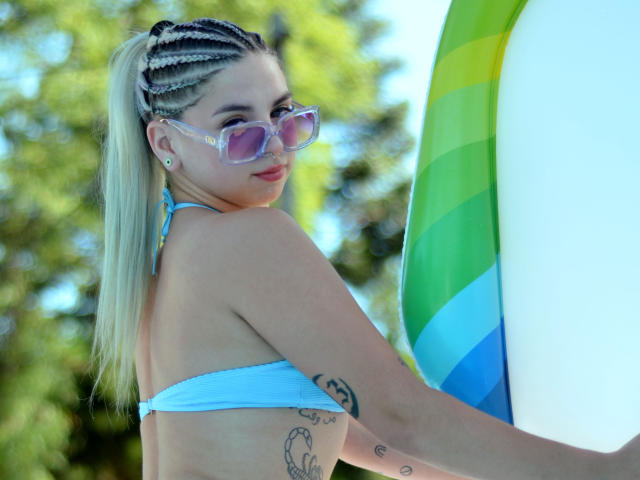KimDenilo with blonde braids, pink sunglasses, blue bikini, holding a green and blue inflatable, showing tattoos on her side, looking back, outdoors, sunny, her ass is visible.