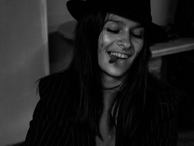 Black-and-white photo of AmelieBlake with long dark hair, wearing a pinstripe suit, black hat, nose ring, and cigarette in mouth, smiling with eyes closed.