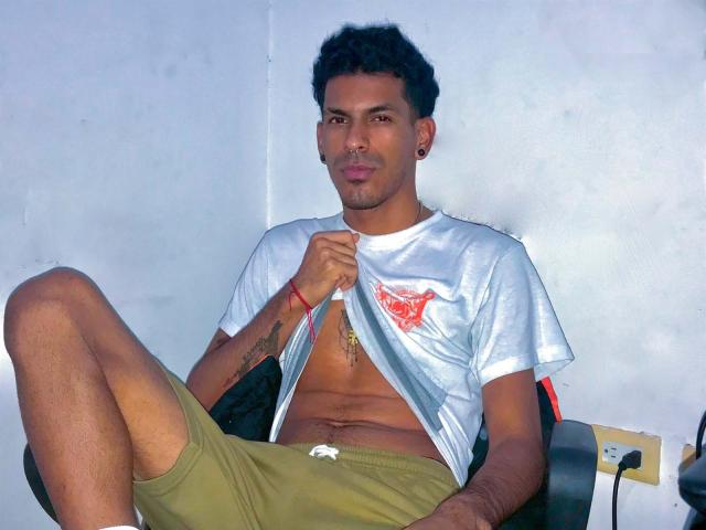 Marco Carter, mit lockigem, schwarzem Haar, sitzt auf einem Stuhl und hebt sein weißes Hemd, um seine trainierten Bauchmuskeln zu zeigen. Er trägt grüne Shorts und ein rotes Armband an seinem linken Handgelenk..