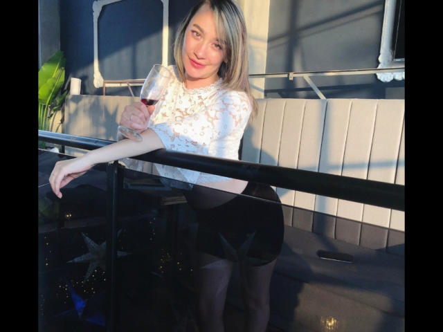 Sarahy Moore mit blonden Haaren, einem weißen Spitzenoberteil und einem schwarzen Rock, hält ein Glas Rotwein, lehnt sich an ein Balkongeländer, lächelt, und ihr Hintern ist leicht angehoben..