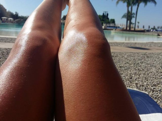 Les jambes lisses et bronzées de JustFuckMeXX, légèrement brillantes à cause de la transpiration, sont écartées sur une plage de galets, révélant leurs cuisses toniques et galbées dans une position détendue..