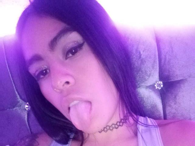 StellaBeckker with dark hair, sticking out her tongue, wearing a nose ring, choker, and sparkly eyeliner, has a cute face and a purple-tinged selfie.