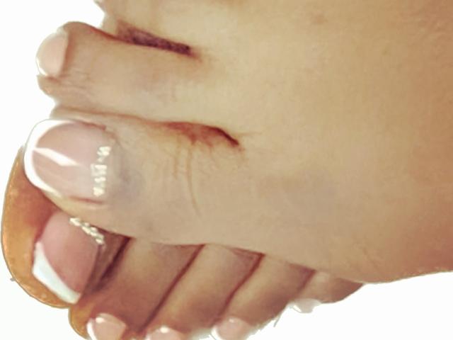 DiosaEbano's finger with neatly manicured, pink and white French tips, gently holding her toes. The skin is light brown with visible texture and slight wrinkles.
