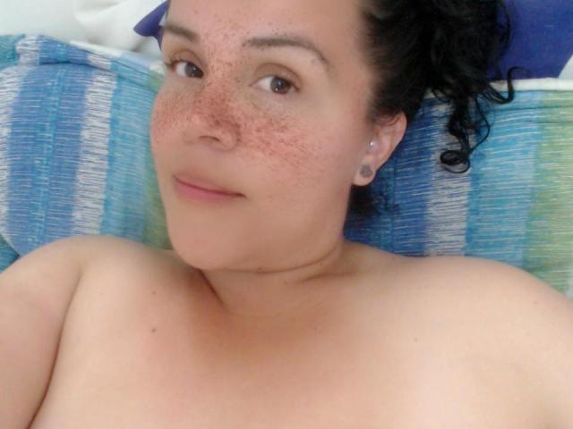 Agatha Miller, senza top, con capelli neri ricci, lentiggini e piccoli orecchini, sorride alla fotocamera, mostrando le spalle scoperte e un leggero rossore sulla pelle..