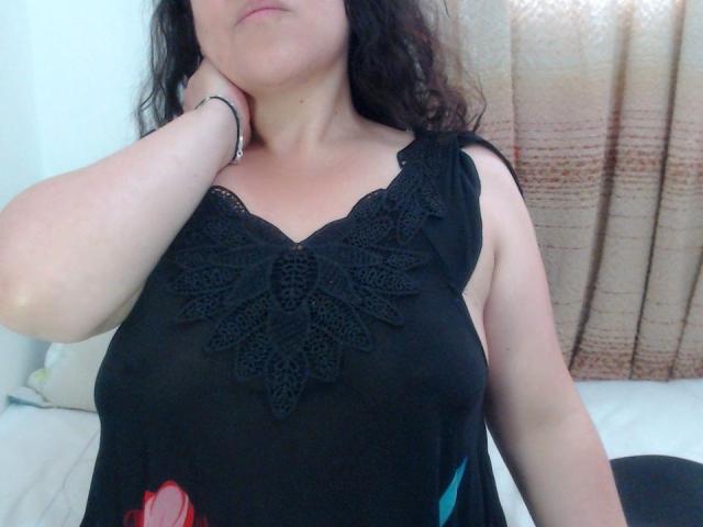 Agatha Miller con una camicetta di pizzo nero che lascia intravedere il seno, braccio alzato, capelli scuri ondulati, pelle chiara, sorriso appena accennato, braccialetto al polso, tenda beige sullo sfondo, letto bianco in primo piano..
