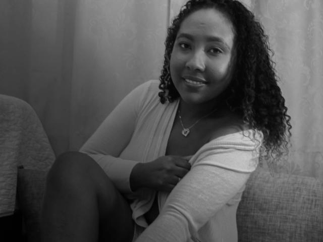 CaroldelaTorre, black woman with curly hair, smiling, wearing a white cardigan, necklace, sitting on a couch, hand on chest, gray background, black-and-white photo.