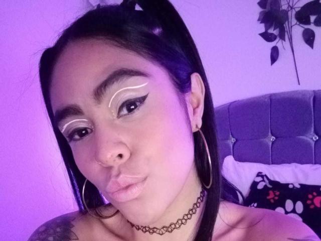 StellaBeckker with black pigtails, white winged eyeliner, gold hoop earrings, nose ring, choker, and puffy pink lips, in purple-lit bedroom, showing off her tattoos.
