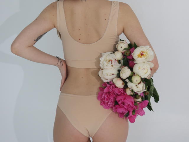 EllenMays mit heller Haut, kleinen Brüsten und einem Tattoo auf ihrem linken Arm, trägt beiges Unterwäsche und hält einen Strauß aus weißen und rosa Blumen vor einer weißen Wand..