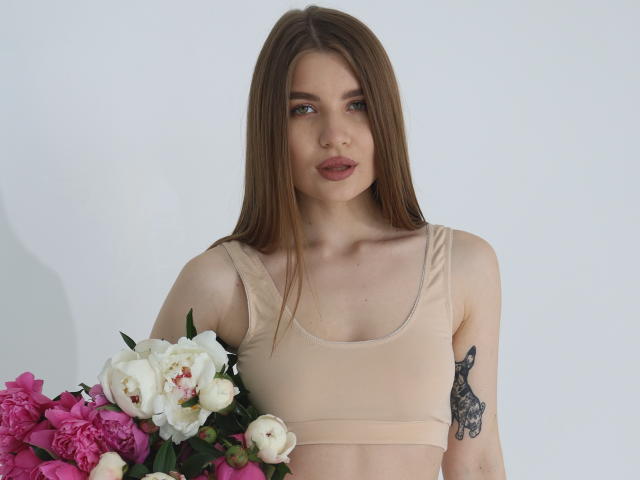 EllenMays with long brown hair, pink lips, wearing a beige sports bra, holding a bouquet of white and pink flowers, has a black tattoo on her right arm.