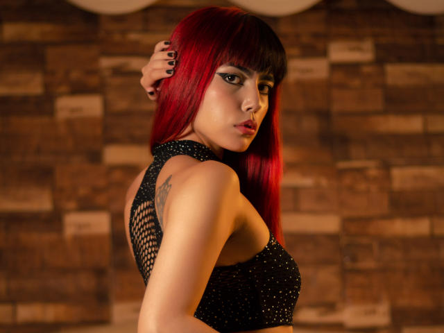 MagicianFoster, avec des cheveux roux, une robe en filet noir, regardant par-dessus son épaule, une main dans les cheveux, montrant un tatouage sur le haut du dos, des ongles peints, sexy, confiante, éclairage chaleureux, maquillage subtil, attirante..