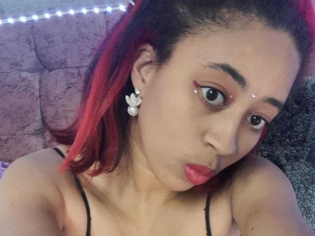 VioletaSteel, com cabelo tingido de vermelho, usando tiras pretas, delineador vermelho, brincos de pérola e batom vermelho, fazendo um leve biquinho, mostrando seu nariz e parte superior do peito. Seus seios estão parcialmente visíveis..