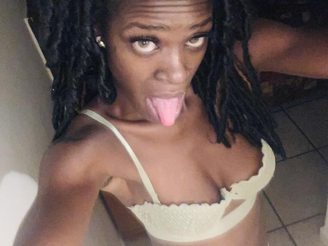 PrettyPrisca, à la peau foncée, avec des dreadlocks, un soutien-gorge vert, la langue rose qui dépasse, regardant vers le haut avec une expression espiègle, montrant sa petite poitrine, dans une pièce faiblement éclairée..