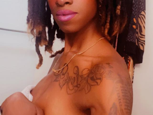 PrettyPrisca, nue, peau brune, rouge à lèvres rose, dreadlocks, collier en or, tatouage sur la poitrine, tatouage sur l'épaule droite, bras croisés, sourire discret, seins de taille moyenne, mamelon visible..