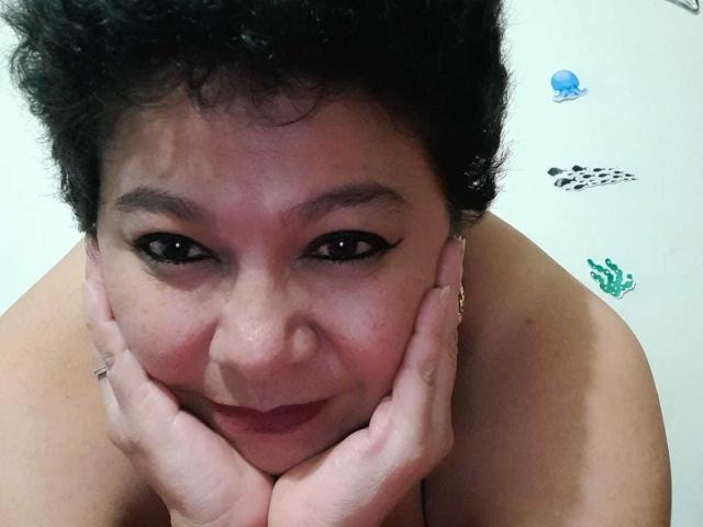 Angellovelly's face, with curly black hair, holding her cheeks, dark eyeliner, and pink lipstick, has her hands under her chin, showing her bare shoulders and a gold ring.