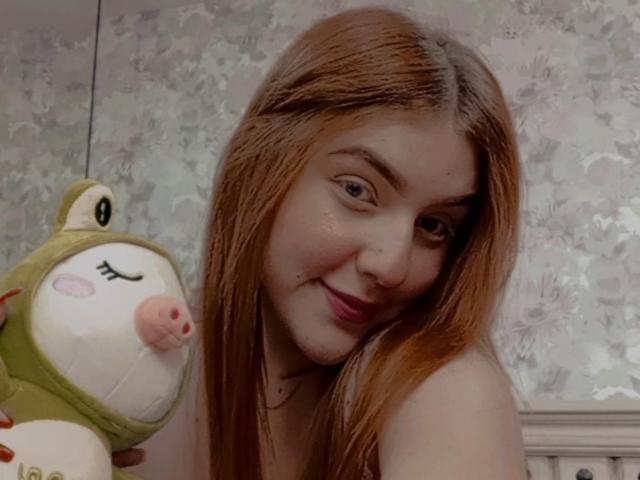 MelisaLee mit langen, braunen Haaren, lächelnd, hält ein grünes Stoffschwein mit rosa Nase, weißem Gesicht und geschlossenen Augen. Ihre Brüste sind teilweise sichtbar. Weiches Licht..