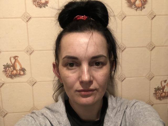 MilashkaLove hat einen Dutt mit einem roten Haargummi, blasse Haut, schwarzes Haar, braune Augen und ein leichtes Lächeln und trägt ein graues Hemd vor einer gefliesten Wand..