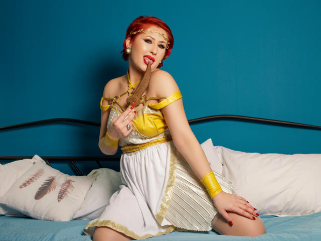 IvyFox con capelli rossi, una toga gialla e bianca, accessori dorati e rossetto rosso, che succhia un bastoncino di legno, inginocchiata su un letto con cuscini bianchi, con una parete blu..