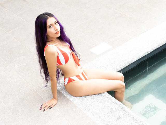 JuliaJobs with long purple hair, wearing an orange and white striped bikini, sitting on a pool edge, showing her ass, with her legs in the water.