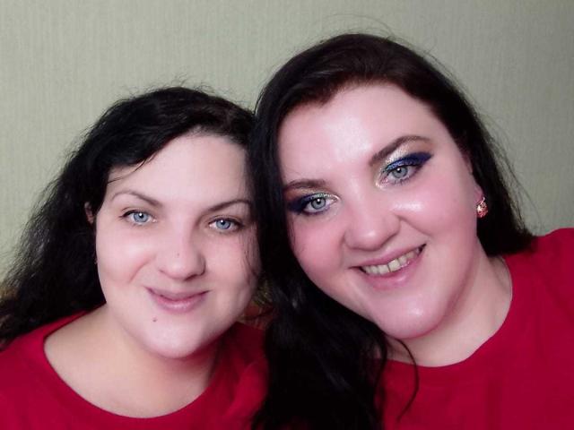 Deux femmes nommées Taisia, avec des cheveux noirs, des yeux bleus et des chemises rouges, souriant à la caméra. La Taisia à droite porte un eye-liner bleu bien visible. Les seins ne sont pas visibles, l'attention est portée sur les visages..