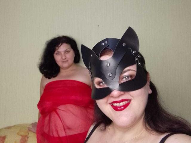 TaisiaW, com máscara de gato preta, batom vermelho e vestido vermelho transparente, exibindo seus seios, senta-se ao lado de TaisiaW, com cabelo preto encaracolado, vestido vermelho e nádegas à mostra..