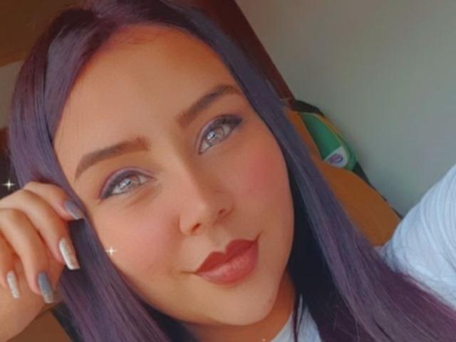 SexyLlianne met paars haar, blauwe ogen en rode lippenstift, die lichtjes glimlacht, met haar hand op haar hoofd, witte nagels, zachte make-up, schattig en sexy, lichte huid, subtiele glans op de wang..