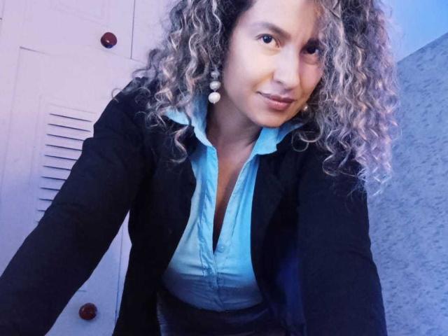 EroticSasha with curly blonde hair, blue shirt, black jacket, and white earrings, leaning forward, smiling, showing a cute, slightly playful expression, tits visible under shirt.