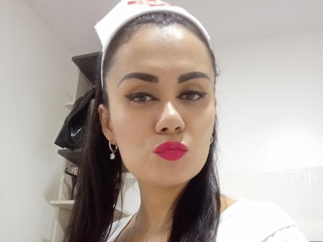 XBellaEvaX with dark straight hair, wearing a nurse cap and white shirt, has pink lipstick, black eyeliner, and hoop earrings, looking at the camera with a slightly pouty expression.