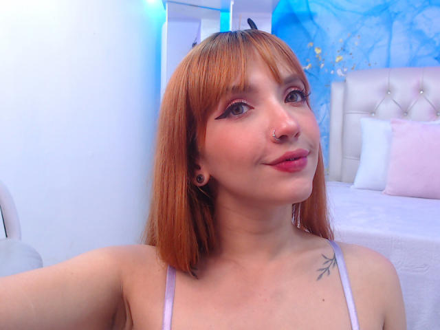 MelissaSkelton with orange hair, nose ring, black ear plugs, small tattoo on right shoulder, smiling, wearing a pinkish-gray strap top, bed with white and pink pillows in background.