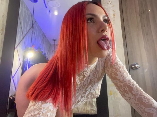 ValeryHarllow, con i capelli rossi, la lingua fuori, indossa una camicetta bianca in pizzo, mostrando il suo sedere scoperto. I suoi lunghi capelli rossi contrastano con la sua camicetta in pizzo e i suoi occhi scuri..