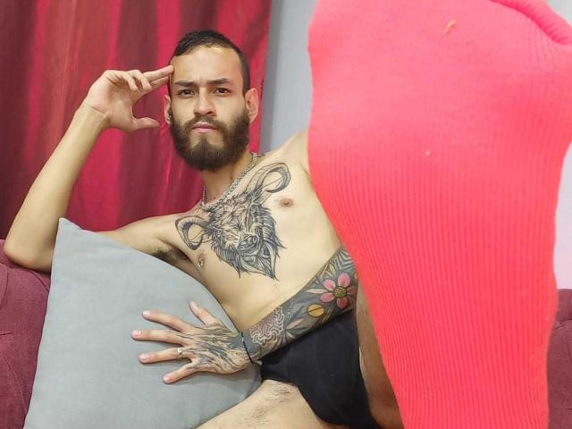 AndersX, con barba, un tatuaje en el pecho de la cabeza de un carnero, ropa interior negra, tatuajes en los brazos y un calcetín rojo en primer plano, sentado en un sofá rojo..