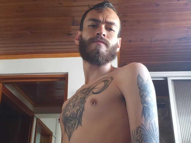 AndersX met een baard, tatoeages op de borst en arm, kort donker haar, die naar beneden kijkt met een lichte grijns, zonder shirt, houten plafond, en een deur op de achtergrond..