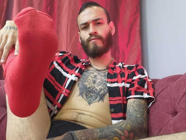 AndersX, avec une chaussette rouge à son pied droit, portant une chemise à carreaux rouge et noir, avec des tatouages sur la poitrine et le bras, une barbe et des cheveux courts, assis sur un canapé rose..
