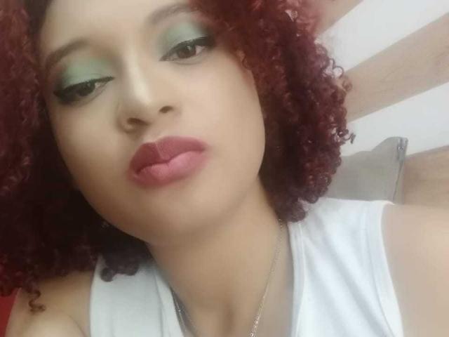 VioletaSwitch has curly red hair, green eyeshadow, and pink lips. She's wearing a white top and a silver necklace, with a neutral expression. Her tits are partially visible.
