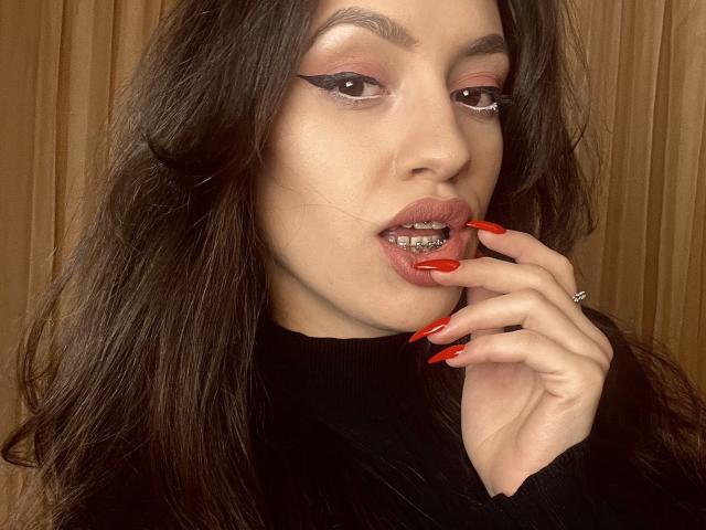 VievienneClemente with dark hair, pink eyeshadow, and red lipstick, holding her finger to her parted lips with red nails, showing metal braces. She wears a black sweater.