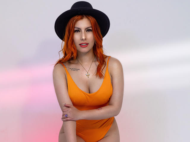 AnnyCraffort with red hair, black hat, orange one-piece, showing sideboob, tattoo on upper arm, looking back, red lipstick, white wall, soft pink and purple light.