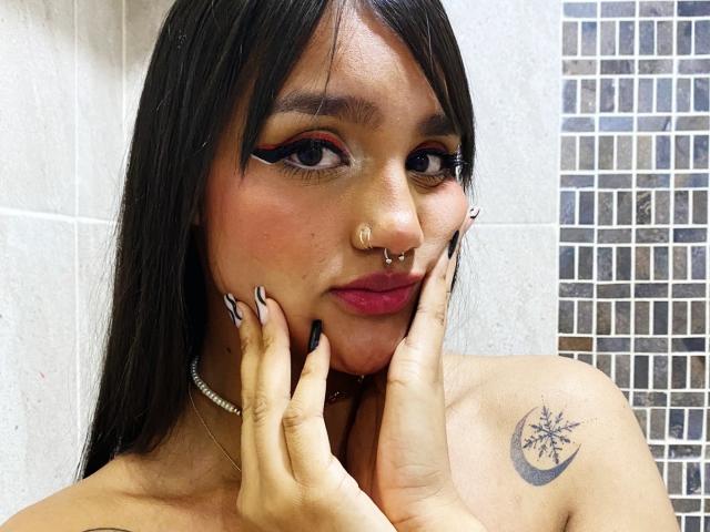 Gaby Durand, avec des cheveux noirs, un trait d'eye-liner audacieux, du rouge à lèvres rose, un anneau au nez, un piercing au septum, un collier ras du cou, un tatouage sur l'épaule droite, les mains sur le visage, des ongles longs, regardant la caméra..