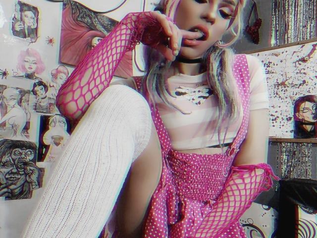 LucyVendeta, com luvas de rede rosa, marias-chiquinhas rosas com bolinhas, blusa listrada de branco, colar de corrente rosa e meias brancas até a coxa, chupando o dedo, mostrando sua vulva com bolinhas rosas..
