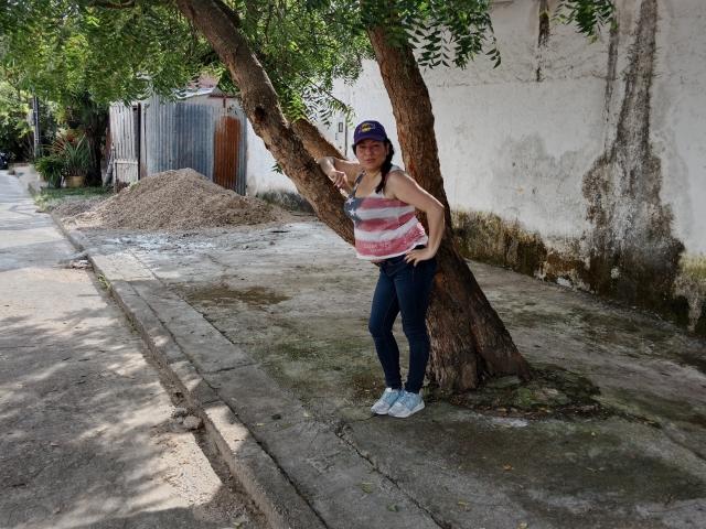 SakotoDika lehnt an einen Baum, trägt ein pinkgestreiftes Tanktop, dunkle Jeans und eine blaue Mütze, hat die Hände in die Hüften gestemmt und lächelt entspannt. Ihre Brüste sind klein..