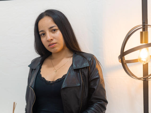 Gianna Russell, con cabello largo y negro, vestida con una chaqueta de cuero negra y una blusa negra con encaje, está de pie cerca de una lámpara esférica encendida. Lleva un pequeño collar con una cruz..