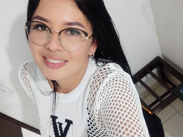 AshleyKatelyn, con cabello negro, gafas y un suéter blanco de malla que deja ver sus senos, sonriendo a la cámara. Lleva lápiz labial rosa y pequeños aretes..