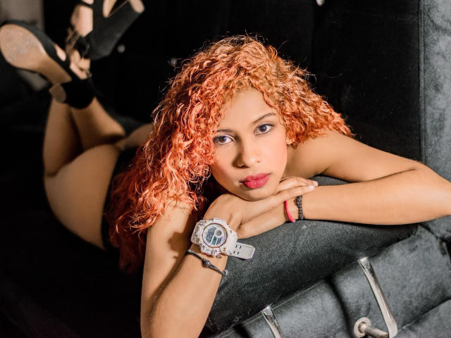 Ariana, eine attraktive Frau mit lockigem, orangefarbenem Haar, liegt auf einer schwarzen Couch, trägt schwarze High Heels, eine Armbanduhr und ein rotes Armband, blickt in die Kamera, hat die Hände auf der Couch und den Po leicht angehoben..