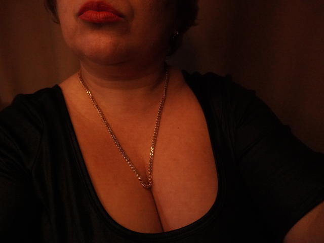 DearLola's chest with large, soft tits, wearing a black shirt and gold chain necklace, has red lips, and is partially lit in dim, warm lighting.
