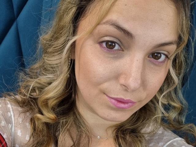MissAnalis a les cheveux blonds et bouclés, un eye-liner rose, du rouge à lèvres rose et un petit nez. Elle porte un haut blanc transparent avec une touche de rouge, et un collier délicat..