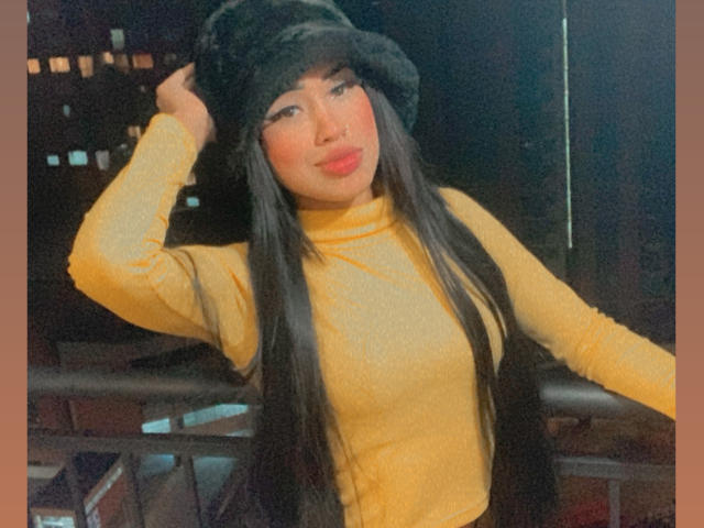 HottsTuffx69 tiene el pelo largo y negro, lleva un cuello alto amarillo, un gorro de piel negra y un chaleco negro. Está haciendo pucheros con lápiz labial rosa, y tiene la mano derecha dentro del gorro. Es de noche..