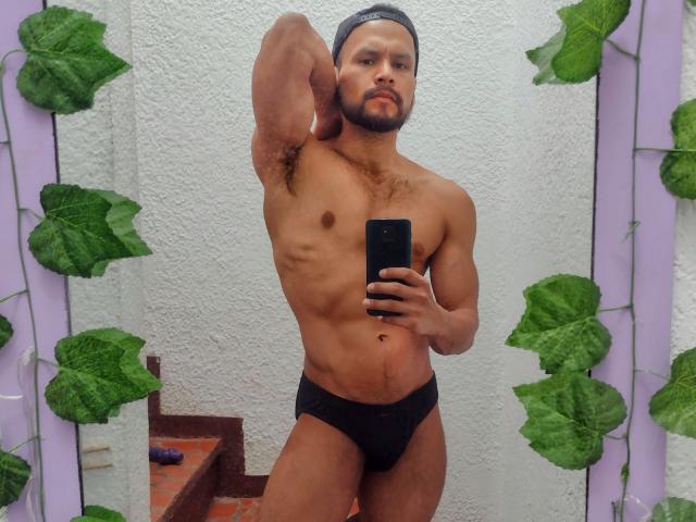 Un hombre musculoso, de piel morena, con ropa interior negra y un gorro, sosteniendo un teléfono, mostrando un pecho fuerte y cubierto de pelo, y posando con confianza y sensualidad en un selfie frente a un espejo..