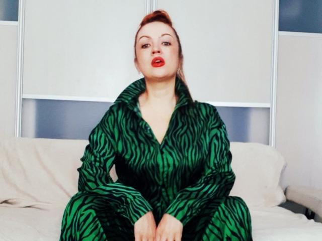 KiraXTender avec du rouge à lèvres rouge, une robe verte à rayures zébrées, assise sur un canapé blanc, les jambes croisées, les mains sur les genoux, regardant la caméra, cheveux bruns attachés en chignon, nez légèrement retroussé..