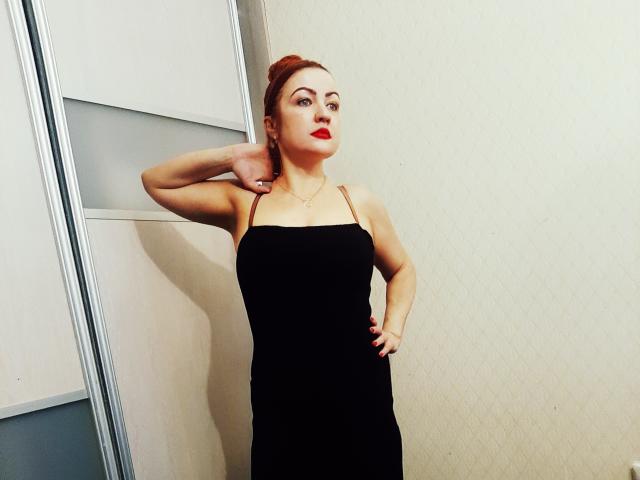 KiraXTender con cabello rojo, vestida de negro, de pie contra una pared, con una mano en el cuello y la otra en la cadera, con lápiz labial rojo, piel pálida, mirando a la cámara..