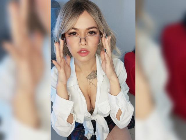 Daria Dawson mit blonden Haaren, Brille und einem weißen Hemd, das offen getragen wird, wobei die Brust betont wird, Hände in der Nähe ihres Gesichts, die in einen Spiegel schaut und dabei ihre Brüste und ein Brust-Tattoo zeigt..