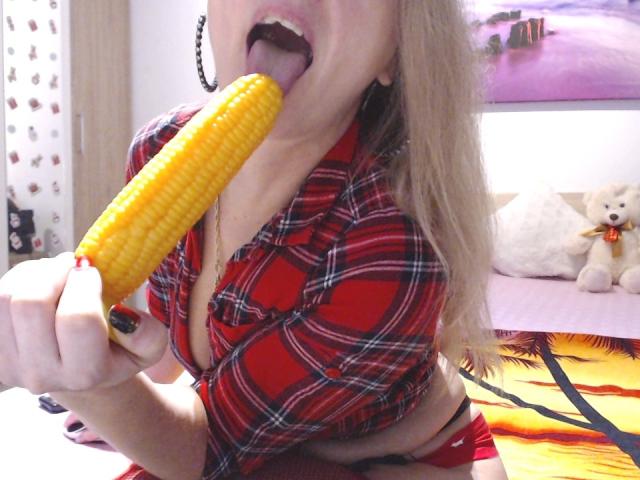 AngryGirl69 with blonde hair, red plaid shirt, and black lipstick, licking a yellow corn on the cob, showing cleavage and black nails, in a bedroom with a teddy bear.